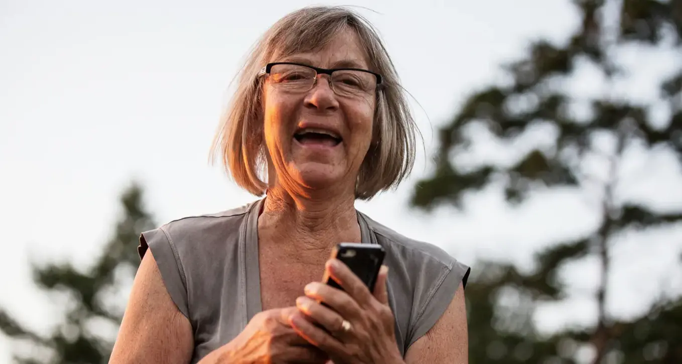 Äldre dam med glasögon skratt och tittar in i kameran med en mobiltelefon i handen